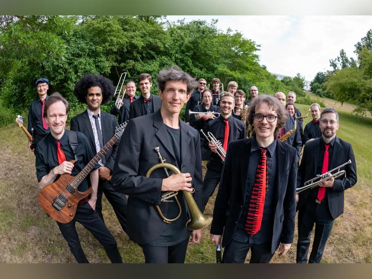 Big Band Mitglieder mit schwarzen Anzügen und roter Krawatte stehen in der Natur mit ihren Instrumenten wie Gitarre, Flügelhorn und Trompete