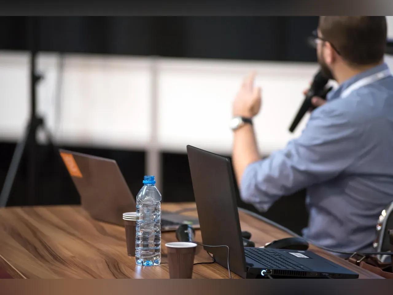 Redner mit Mikrofon hinter einem Tisch mit Laptops und Getränken