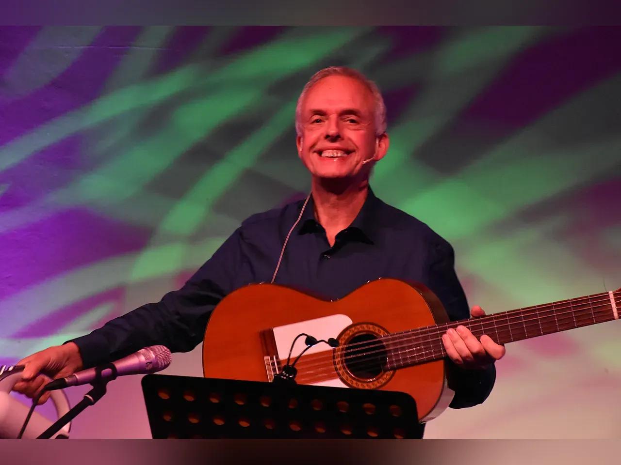 Ein Gitarrenspieler mit grauem Haar lächelt und sitzt auf einer Bühne.