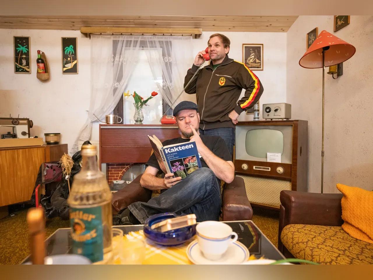 Zwei Autoren sitzen in einer Wohnstube mit DDR-Interieur.