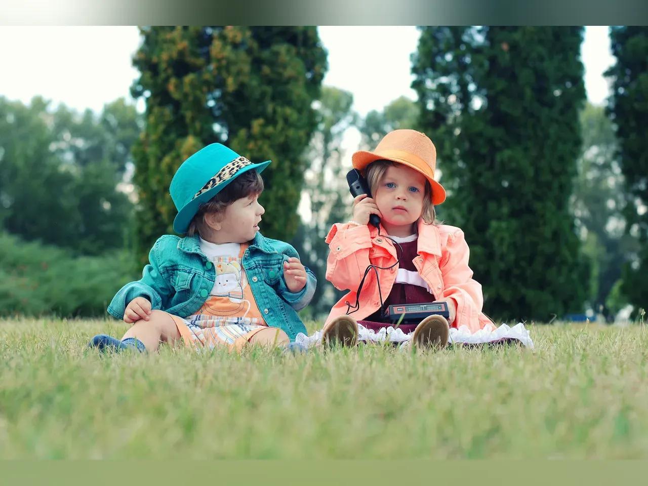 Zwei Kinder in farblich passenden Jäckchen und Hüten sitzen auf einer Wiese, das rechte Kind hat ein Telefon am Ohr.