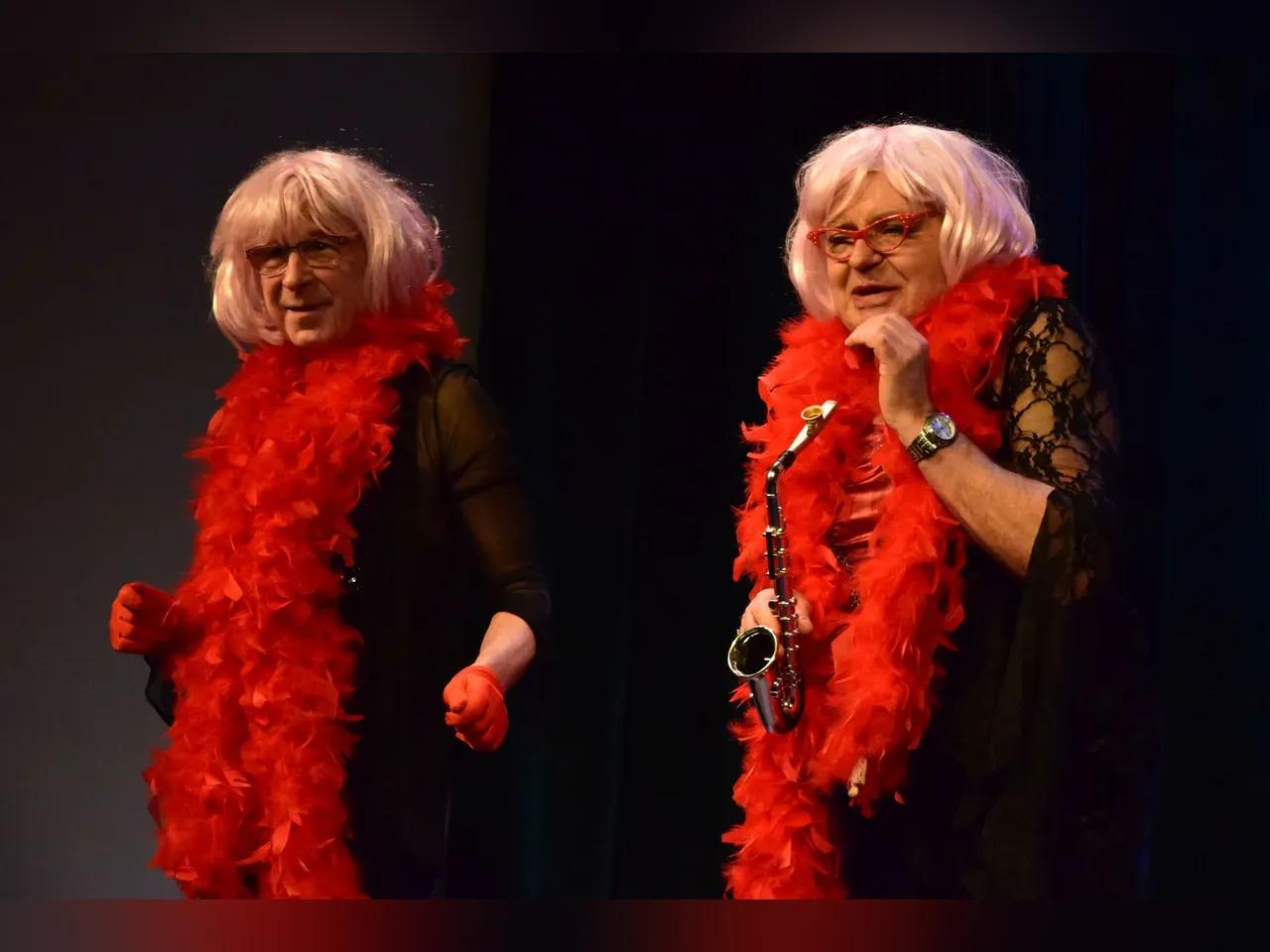 Zwei Männer auf der Bühne in Frauenkleidern und Perrücken, einer von Ihnen spielt Saxophon.