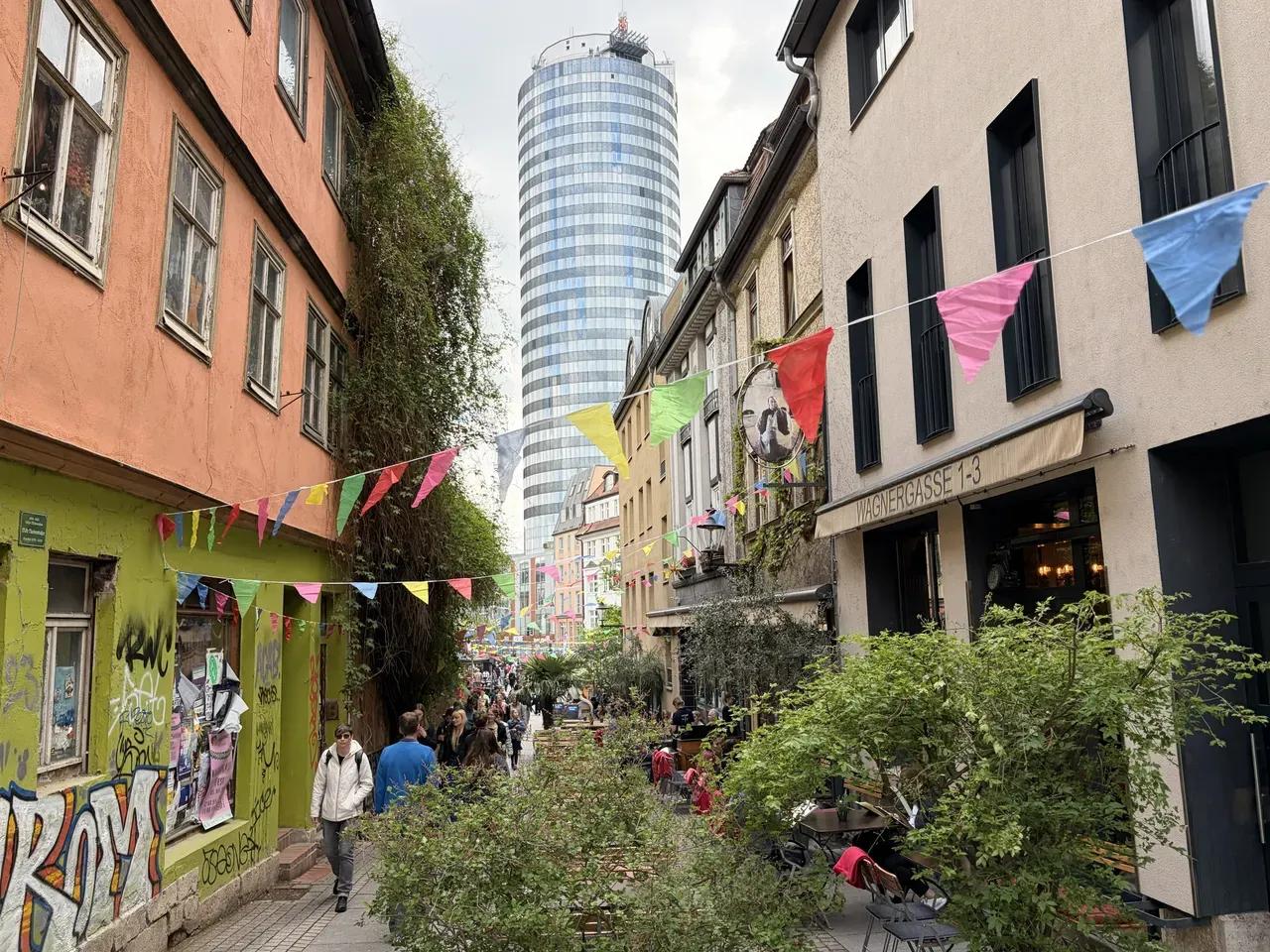 Belebte Wagnergasse in Jena mit bunten Wimpelketten, Fußgänger:innen und dem JenTower im Hintergrund.