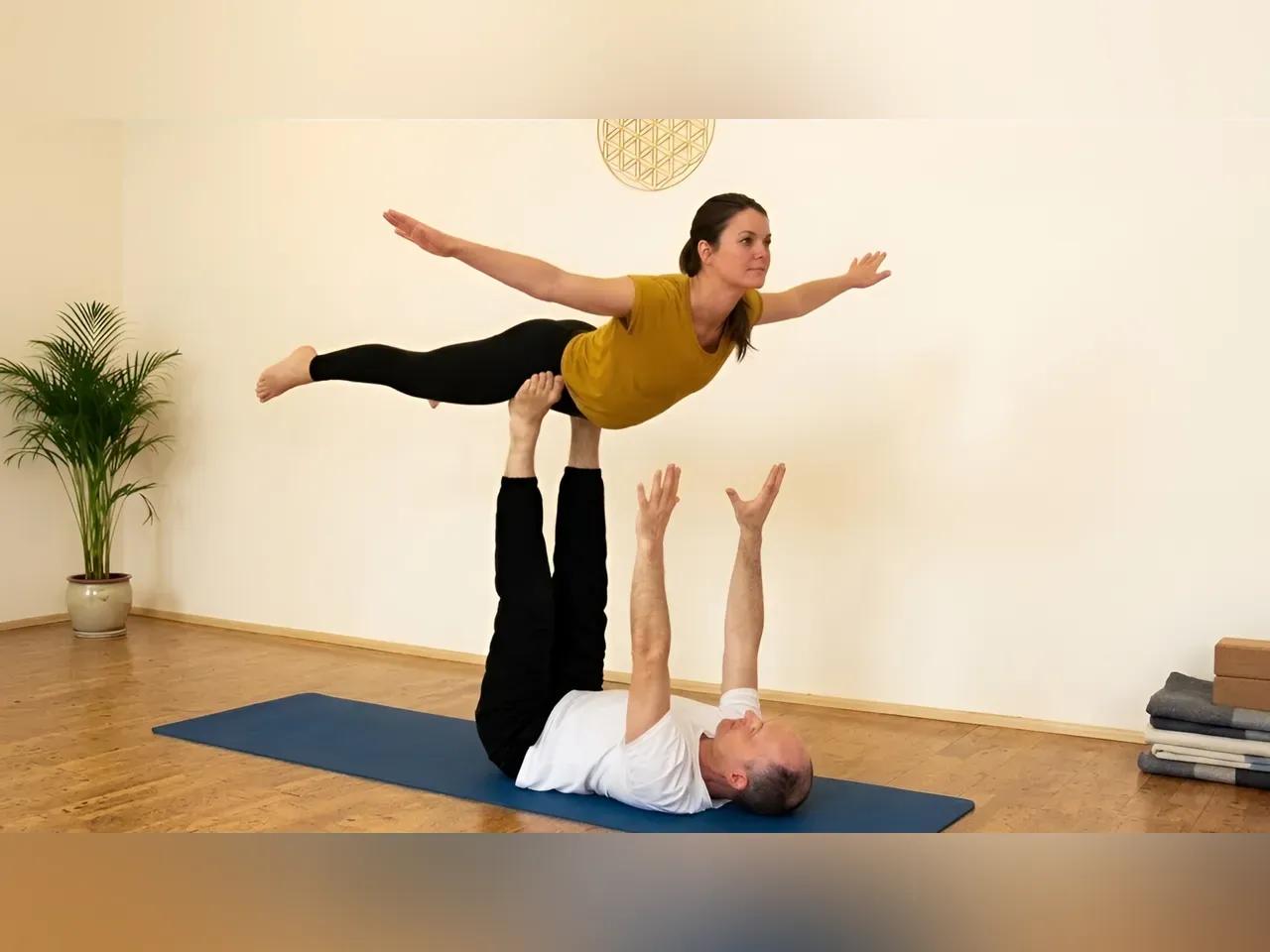 Ein Mann und eine Frau beim Acrobatic Yoga: Er liegt auf dem Rücken, sie auf seinen Füßen in der Luft